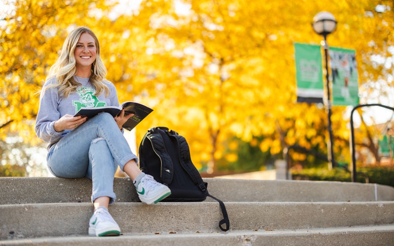 A student sits on the plaza in front of fall trees on Marshall University's campus