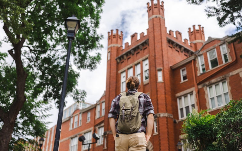 A student wearing a backpack looks up at Marshall University's Old Main from the sidewalk