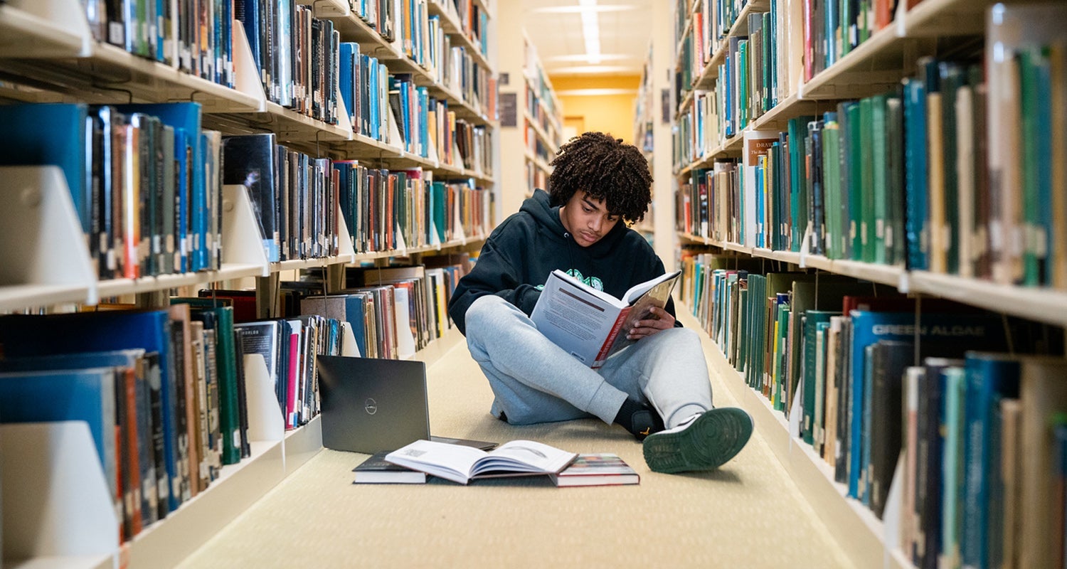 A student sits on the floor of the library with a laptop, surrounded by books.