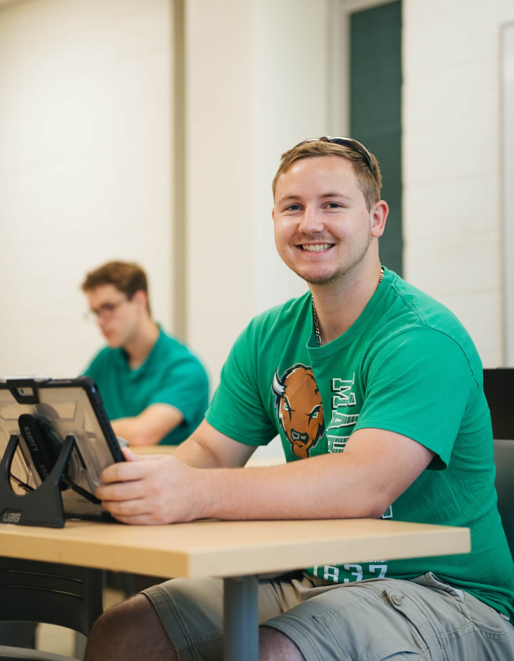 male student sitting at a desk with an iPad smiling at the camera